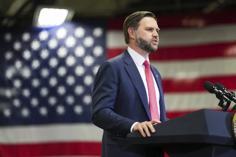 A man in a suit stands at a podium in front of an American flag, delivering a speech.
