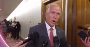 A man in a suit and tie is speaking to reporters in a hallway.