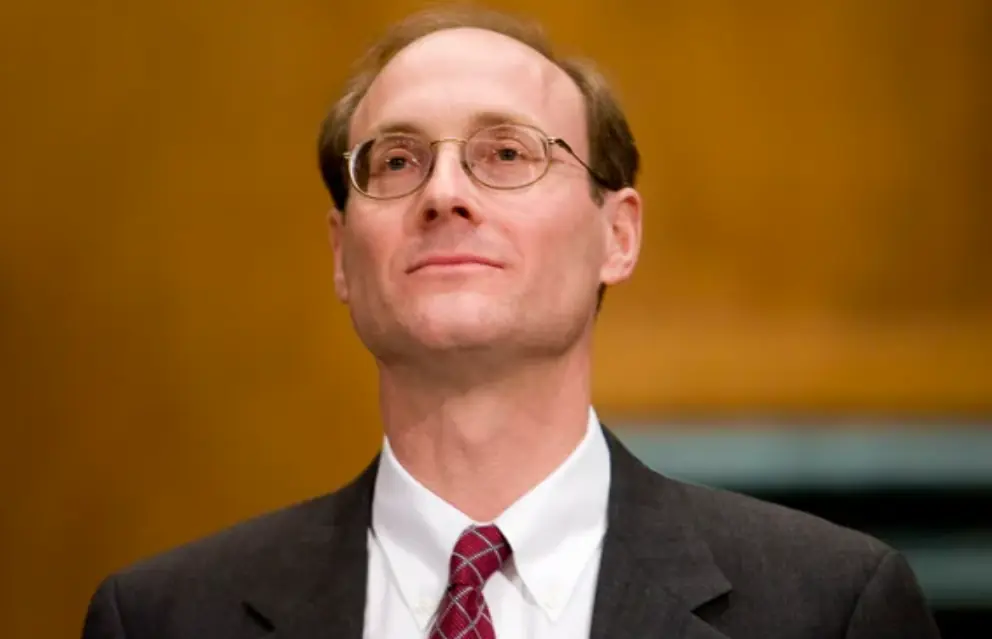 The image shows a man in formal attire, wearing glasses and a suit with a red tie, looking upward with a serious expression.
