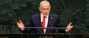 The image shows a man in a suit and red tie speaking at a podium with microphones, likely at a formal international event.