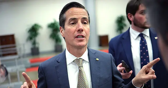 A man in a suit and tie is gesturing with his hands while speaking, with another individual in the background holding a phone.