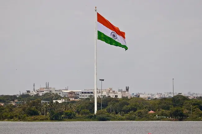 The Indian national flag waves prominently against a backdrop of urban buildings and greenery.