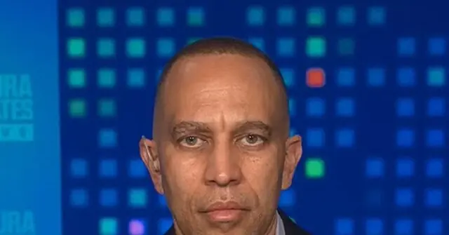 The image shows a person in a news broadcast setting, with a blue background featuring a grid pattern and some text partially visible on the left side.