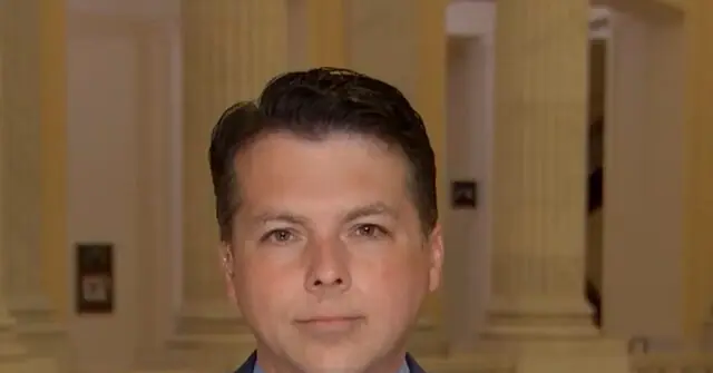 The image shows a person in a formal setting, possibly a government or institutional building, with columns and a door in the background.