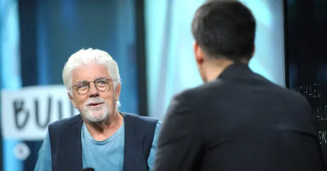 The image shows a man with white hair and a beard, wearing glasses and a blue shirt, engaged in a conversation with another person whose back is to the camera.