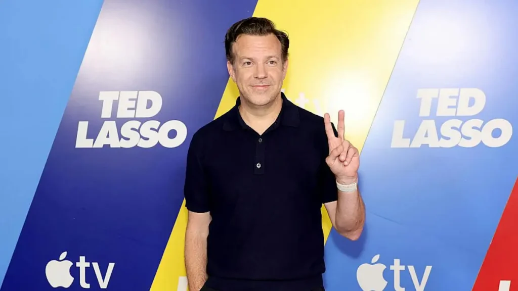 The image shows a person posing in front of a backdrop featuring the "Ted Lasso" logo and the Apple TV+ logo, making a peace sign with their hand.