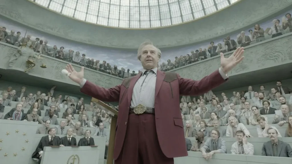 A man in a red suit stands with his arms outstretched in front of a large audience in a grand hall.
