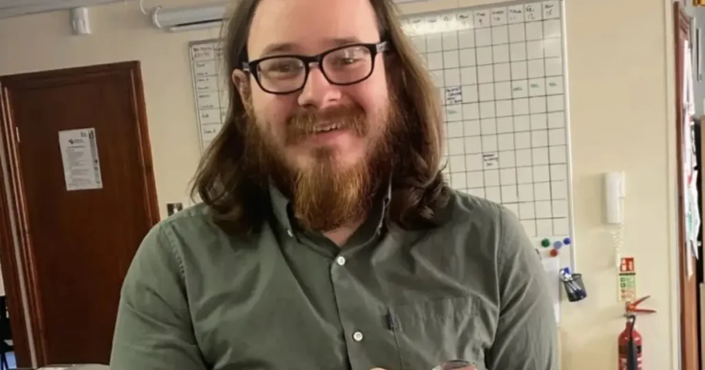 The image shows a person with long hair and a beard, wearing glasses and a green shirt, standing in an office setting with a whiteboard and various office supplies in the background.
