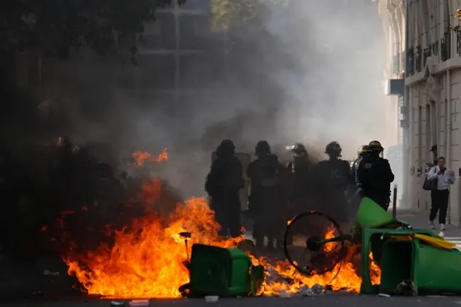 A group of police officers in riot gear stand near a burning barricade during a protest.