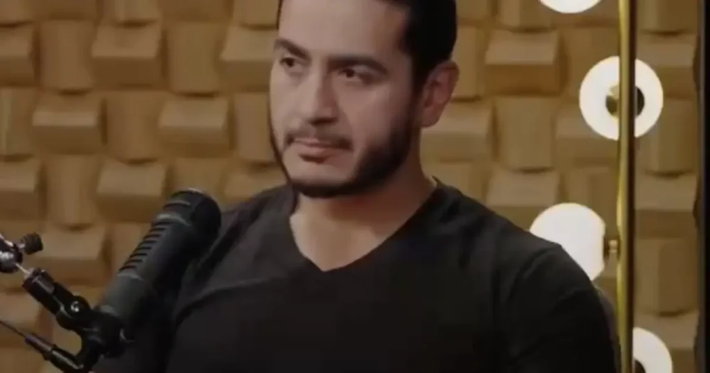 The image shows a person speaking into a microphone in a studio setting with acoustic foam panels in the background.