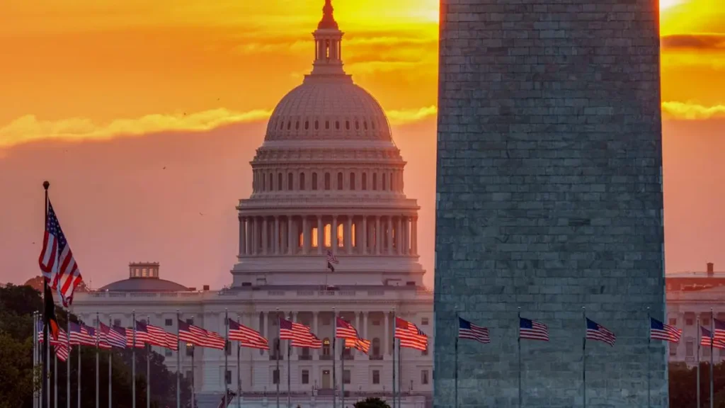The image captures the iconic U.S. Capitol and Washington Monument at sunset, with American flags lining the foreground.