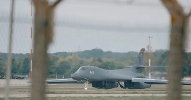 A military aircraft is taxiing on a runway, viewed through a tree-lined perspective.