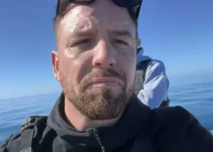 The image shows a person wearing a black jacket and sunglasses, standing outdoors with a clear blue sky and water in the background.