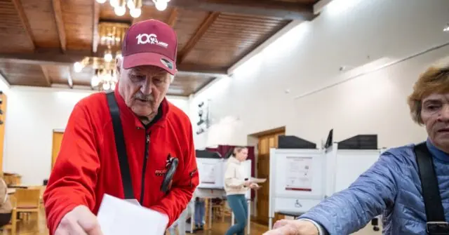 An elderly man wearing a red jacket and a cap is seen at a polling station, holding a piece of paper, while another person in a blue jacket is assisting him.