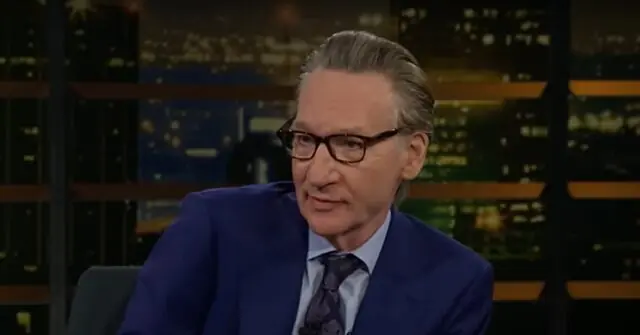 The image shows a man in a blue suit and tie, wearing glasses, seated in a studio setting with a cityscape background.