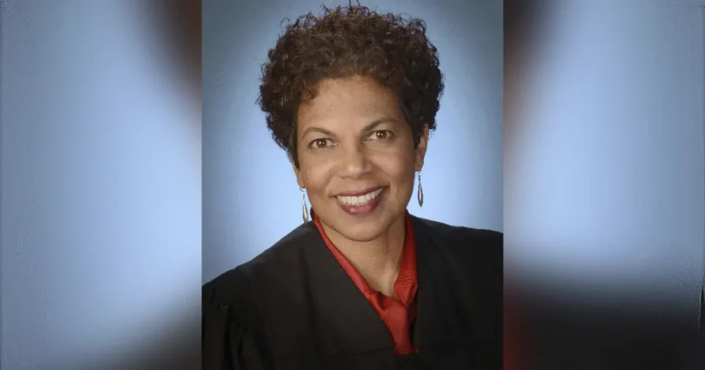 The image shows a professional portrait of a smiling individual wearing a black blazer over a red shirt, set against a soft blue background.