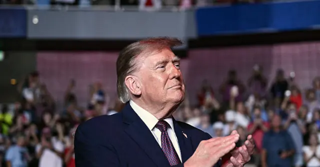 The image shows a man in a suit clapping in front of a crowd, likely at a public event or rally.
