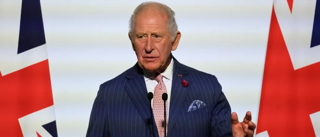 The image shows a man in a formal blue suit and tie, standing at a podium with microphones, with the Union Jack flag in the background, suggesting a formal or official event.