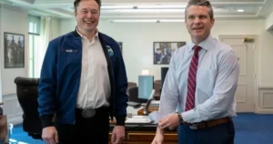 Two men are standing in an office setting, smiling and holding papers, with one wearing a jacket labeled "Musk."