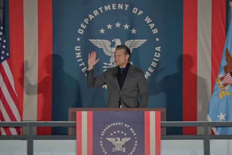 A man stands at a podium in front of a backdrop featuring the Department of War seal and American flags, raising his hand as if making a pledge or taking an oath.
