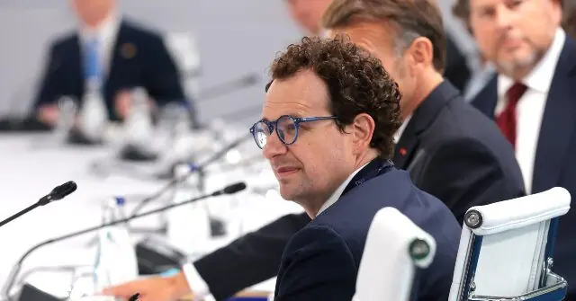A man in a suit and glasses is seated at a conference table with microphones, appearing to listen attentively during a formal meeting.