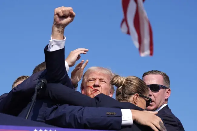 The image shows a political figure raising his fist in a gesture of defiance or solidarity, with an American flag visible in the background.
