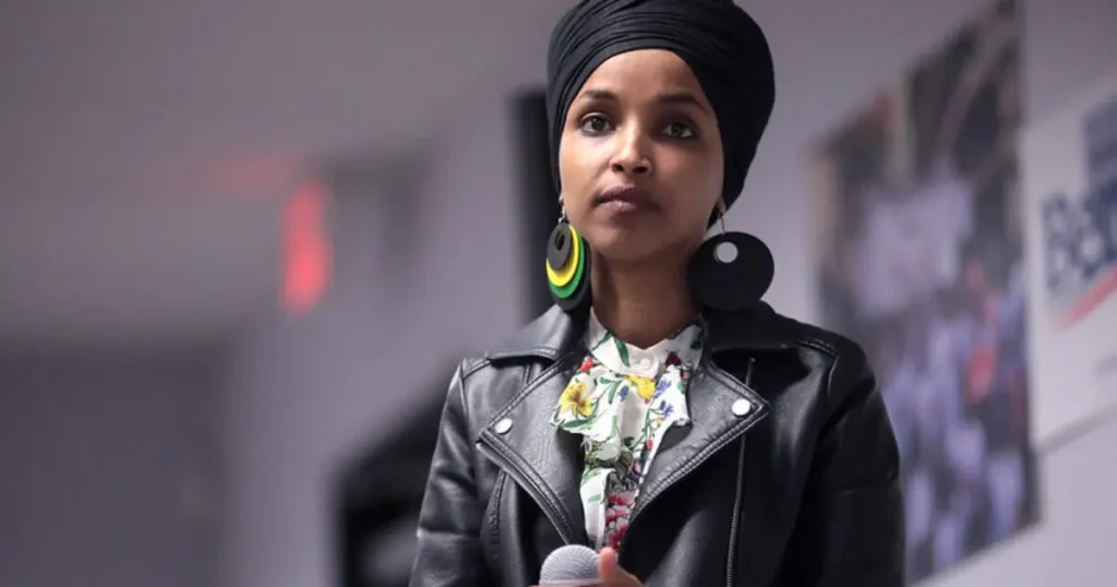A woman wearing a black turban and a leather jacket is speaking into a microphone, with a colorful floral scarf and large earrings.