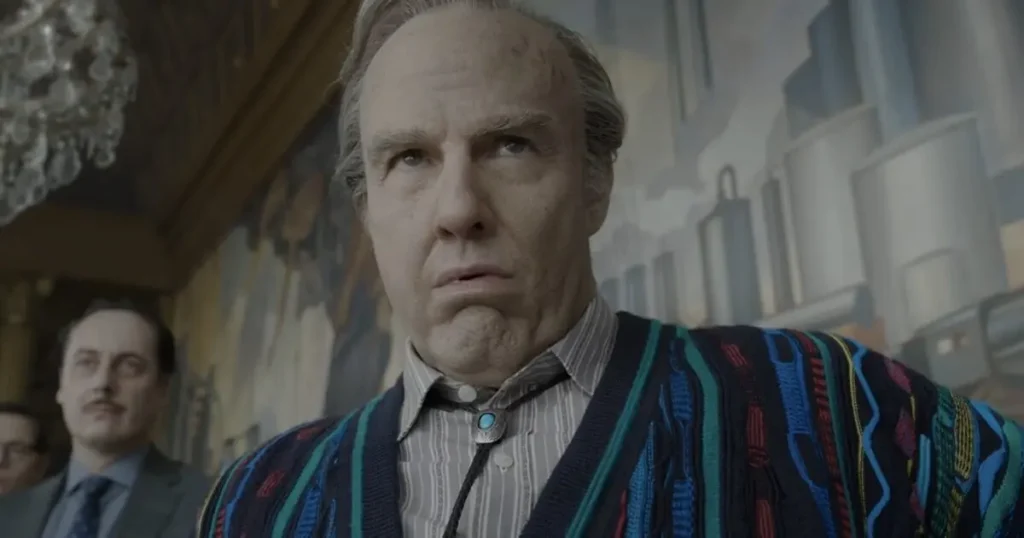 The image depicts a man in a colorful striped sweater and a bow tie, standing in a room with a chandelier and a mural in the background.