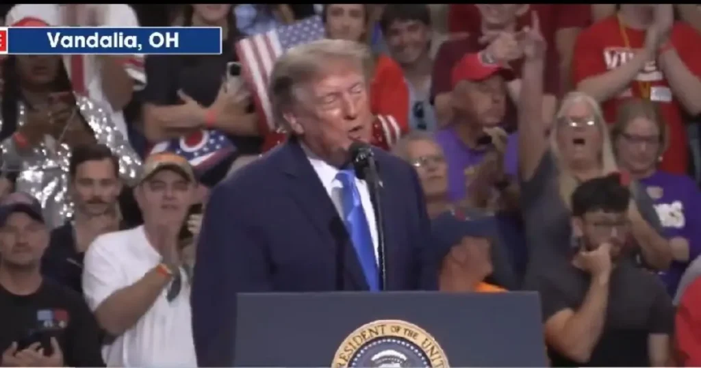 The image shows a political rally in Vandalia, OH, with a speaker at a podium and a crowd of supporters cheering and clapping.