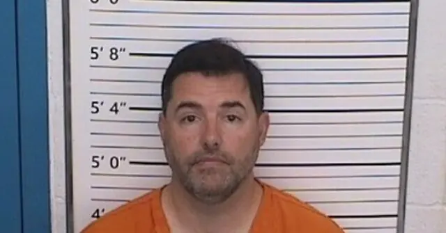 The image shows a mugshot of a man in an orange jumpsuit standing in front of a height chart.
