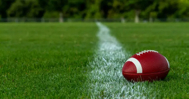 A red American football rests on a grassy field marked with a white line.