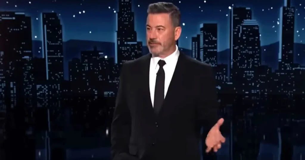 The image shows a man in a black suit and tie standing on a stage with a cityscape background, likely during a talk show or similar event.