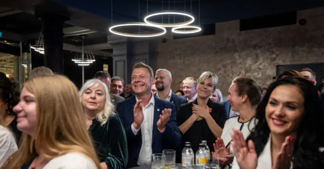 A group of people are clapping and smiling in a modern indoor setting with circular pendant lights.