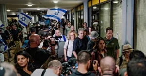 A crowd of people, some holding Israeli flags, is gathered in a public space, with media personnel and photographers present.