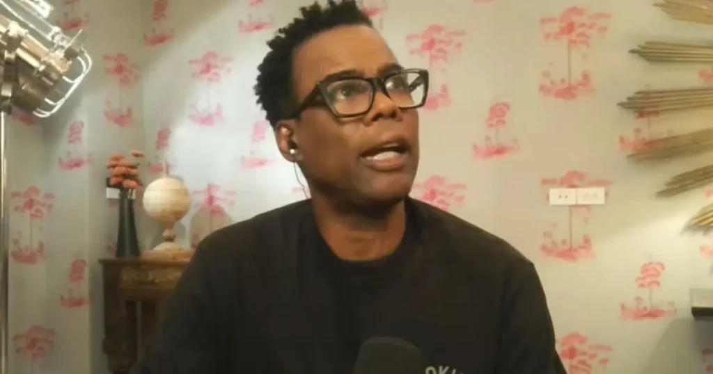 The image shows a person wearing glasses and a black shirt, speaking into a microphone in front of a wall with a red and white pattern.