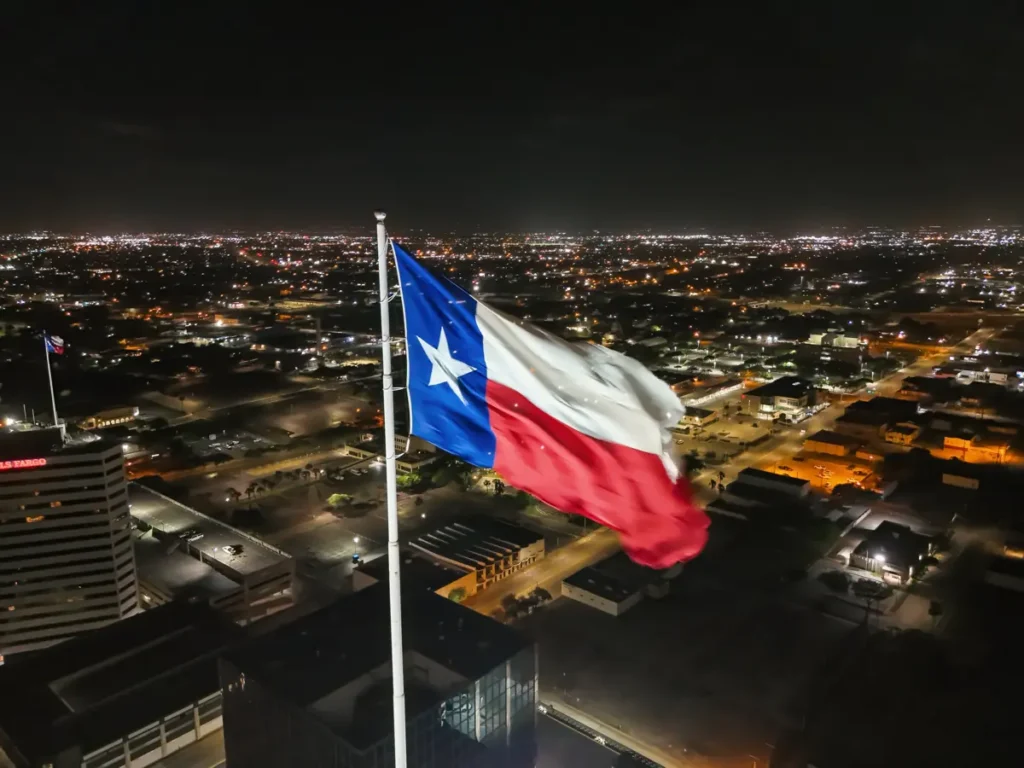The Texas state flag waves prominently against the backdrop of a cityscape illuminated at night.