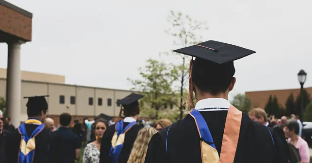 A graduate in a black gown and cap walks through a crowd of fellow graduates and attendees at a graduation ceremony.