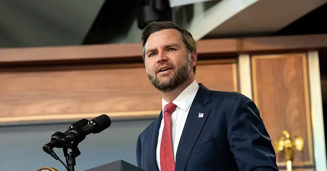 A man in a suit and tie stands at a podium, speaking into a microphone in a formal setting.