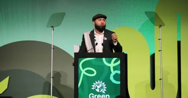 A man stands at a podium with a "Green" sign, speaking at an event with a green-themed backdrop.