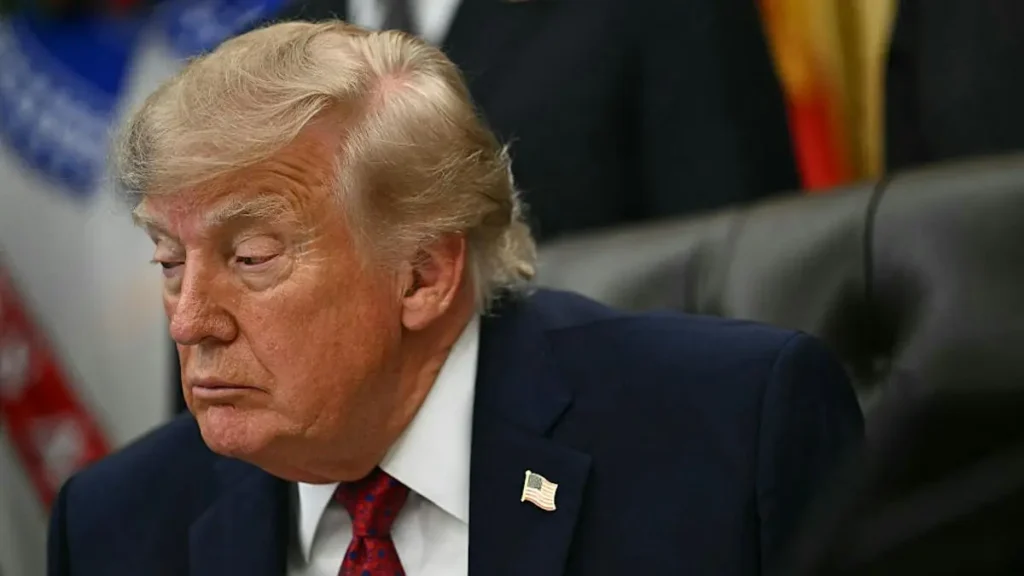 The image shows a person in a suit with an American flag pin, seated in a formal setting with flags in the background.