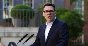 A man in a suit stands at a podium with microphones, speaking outdoors in front of a building.