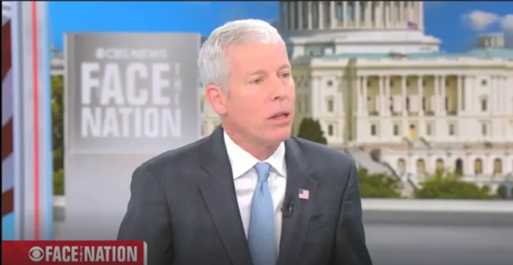 The image shows a man in a suit and tie on a news program, with the Capitol building in the background and the "Face the Nation" logo visible.