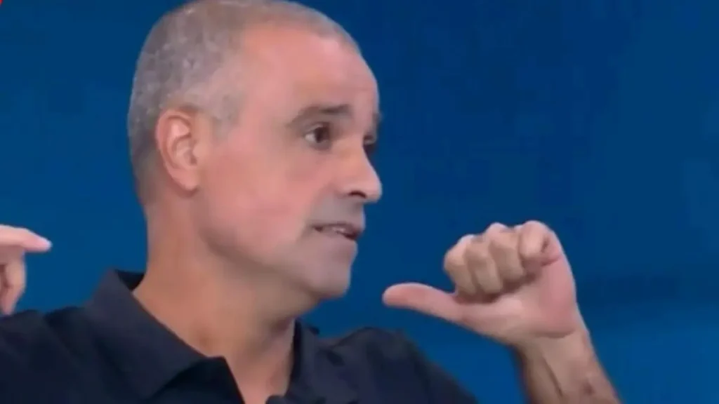 The image shows a man gesturing with his hands while speaking, set against a blue background.