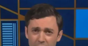 The image shows a close-up of a news anchor against a blue and yellow background.