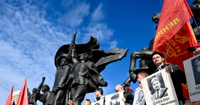 The photo depicts a group of people holding red flags and portraits, standing in front of a large statue, possibly during a commemorative or political event.