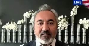 The image shows a man speaking in a live broadcast, with a backdrop of silver vases and white flowers.