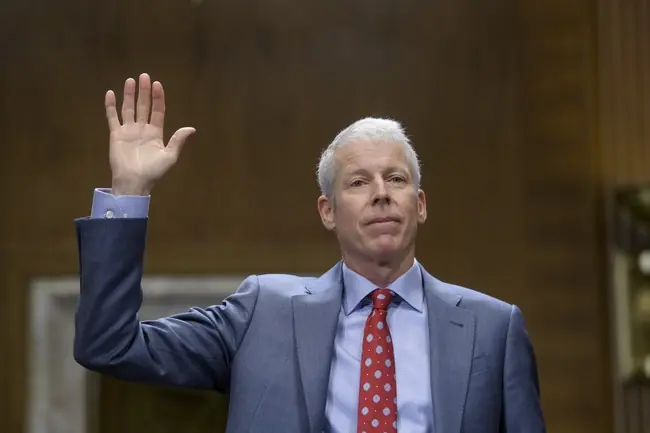 A man in a suit raises his hand, possibly taking an oath or making a gesture.