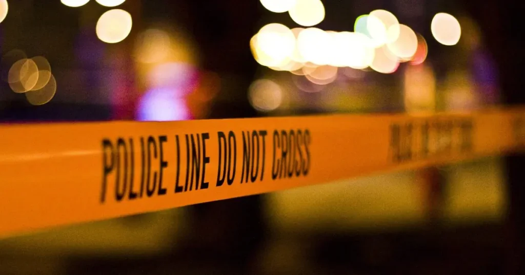 The image shows a "POLICE LINE DO NOT CROSS" tape at a crime scene with blurred lights in the background.