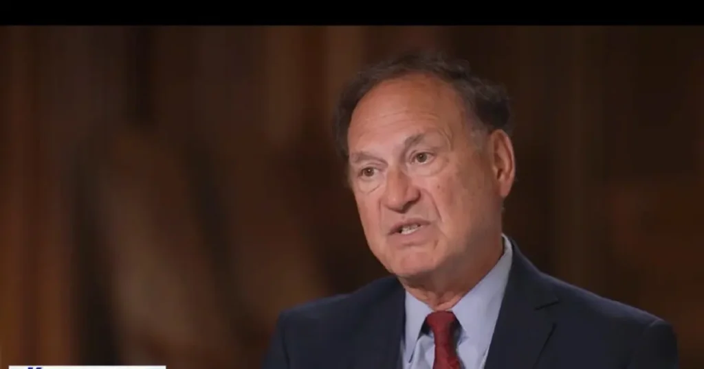 The image shows a man in a suit and tie, speaking in what appears to be a formal or professional setting.