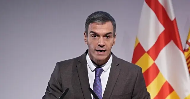 The image shows a man in a suit and tie speaking at a podium with a Catalan flag in the background.